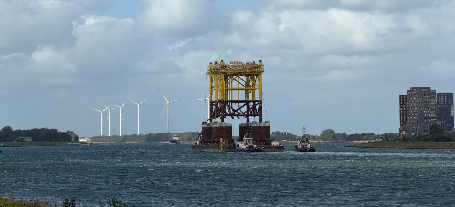 Gele offshore-constructie op een ponton, begeleid door sleepboten, met windturbines op de achtergrond.